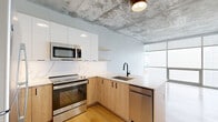 This thoughtfully designed 2-bedroom, 1-bathroom unit spans a single floor and showcases a compelling blend of industrial and contemporary aesthetics. Throughout the space, light wood flooring and exposed concrete ceilings create a striking visual contrast that defines the unit's character.

The open-concept living area seamlessly connects the kitchen, dining, and living spaces. The kitchen is a standout feature, equipped with stainless steel appliances including a range, microwave, refrigerator, and dishwasher. Warm natural wood cabinetry is paired with crisp white upper cabinets, a marble-look quartz backsplash, and matching quartz countertops. Matte black fixtures add a modern, cohesive touch. Floor-to-ceiling windows flood the living area with abundant natural light and offer views of the surrounding exterior.

The bedrooms are well-proportioned, with the primary bedroom featuring its own exposed concrete ceiling detail and access to natural light. Three closets throughout the unit provide ample storage.

The full bathroom is finished with a light wood vanity, white quartz countertop, dual undermount sinks, matte black hardware, a glass-enclosed shower, and neutral tile flooring. A dedicated laundry area and central hallway complete the functional layout of this well-appointed urban unit.
