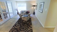 This single-floor space showcases a welcoming living room as its centerpiece. The room is finished with wall-to-wall carpeting in a warm neutral tone, complemented by crisp white baseboards and soft, muted wall colors that create a calm, cohesive atmosphere. A large window along the far wall allows generous natural light to filter into the space, offering a view of greenery outside and enhancing the room's airy, open feel. The living area is anchored by a plush, oversized armchair paired with a tufted ottoman, both positioned atop a bold, swirling patterned area rug in deep browns and blacks that adds a striking visual contrast to the light flooring. Decorative touches include tall floor vases, a slender table lamp, and framed wall art that adds personality to the neutral backdrop. The layout is open and functional, allowing for easy movement throughout the space. Overall, the living area balances comfort and style, offering a relaxed yet put-together environment suited for everyday living.