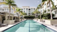 This residential community showcases a striking outdoor amenity area centered around a generously sized swimming pool with clear, turquoise water. The pool is framed by a clean, light-colored pool deck furnished with modern lounge chairs and sun loungers, providing ample space for relaxation. Tall, mature palm trees are planted throughout the surrounding area, lending a tropical, resort-like ambiance to the space. Neatly trimmed hedgerows and verdant landscaping add to the lush, well-maintained aesthetic.

The backdrop is defined by a contemporary multi-story building with a white exterior, featuring large windows and private balconies on each level, offering residents elevated views of the pool and greenery below. The architectural design is clean and modern, with a symmetrical facade that contributes to the overall sense of order and elegance. The open-air setting allows abundant natural light to filter through the palm fronds, creating a bright and inviting atmosphere throughout the day. The combination of tropical landscaping, modern architecture, and resort-caliber amenities gives this community a distinctive and upscale character.