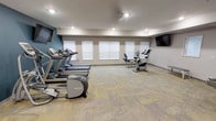 This single-story fitness center offers a clean, functional environment designed to support cardio-focused workouts. The space is outfitted with a row of treadmills and an elliptical machine positioned along one side, complemented by two recumbent stationary bikes on the opposite end. A wall-mounted flat-screen television is positioned for easy viewing during workouts. Natural light filters in through multiple windows fitted with white horizontal blinds, brightening the neutral beige and grey-toned walls. Recessed ceiling lighting provides additional illumination throughout the space. The floor is covered in durable, patterned carpet tiles in muted tones, offering comfort underfoot while maintaining a practical, low-maintenance surface. An accent wall in a muted teal-blue hue adds a subtle visual element to the otherwise neutral color palette. The layout is open and uncluttered, allowing for ease of movement between equipment. The overall ambiance is straightforward and utilitarian, providing a focused environment suited for daily cardio exercise routines.