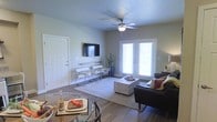 This single-floor residence offers a thoughtfully designed open-concept layout that seamlessly connects the dining and living areas, creating a comfortable and functional living environment. The space features warm hardwood-style flooring throughout, which lends a modern yet inviting aesthetic to the interior.

The living area is the centerpiece of the home, highlighted by a ceiling fan that provides both style and comfort. A wall-mounted flat-screen television anchors one side of the room, while a set of elegant French doors floods the space with natural light and offers a connection to the outdoors. The living room comfortably accommodates a plush sofa, a woven coffee table, and a pair of sleek accent chairs, creating a balanced and welcoming seating arrangement.

Adjacent to the living area, the dining space features a glass-top table with coordinating chairs, ideal for casual meals or entertaining. Neutral-toned walls in a soft green-grey palette complement the overall design, creating a calm and cohesive atmosphere throughout. Decorative touches, including potted greenery, add a fresh, natural element to the interior. The space strikes an ideal balance between practicality and style, making it a comfortable and attractive single-floor home.