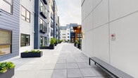 Located at 633B SW 150th St in Burien, Washington, this single-level outdoor amenity space is set within a contemporary multi-story residential community. The courtyard is defined by a clean architectural palette of navy blue horizontal siding and crisp white panel cladding, creating a polished, modern aesthetic throughout.

The expansive paved courtyard serves as the central gathering area, finished with large-format light gray tiles that provide a sleek, durable surface. A standout feature is the oversized outdoor chess set, embedded directly into the patio surface, offering an engaging, interactive element for residents. Flanking the courtyard are generously sized black metal planters filled with lush greenery and shrubs, adding natural texture and softness to the hard architectural lines.

Comfortable outdoor seating clusters are arranged along one side, providing dedicated areas for relaxation and socializing. An outdoor television, housed behind sliding panel shutters, adds an entertainment dimension to the space. Warm wall-mounted sconces illuminate key architectural corners, enhancing the ambiance during evening hours. Sliding glass doors connect ground-floor units directly to the courtyard, seamlessly blending indoor and outdoor living in this well-appointed community setting.