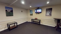 This single-floor space presents a clean, functional layout designed for comfort and utility. The room features classic wainscoting along the lower portion of the walls, painted in a neutral cream tone that lends a polished, professional appearance. Dark charcoal carpeting covers the floor, providing a grounded contrast to the light-colored walls and contributing to a quiet, composed atmosphere.

Recessed ceiling lights provide even, ambient illumination throughout the space. A wall-mounted flat-screen television, supported by a ceiling-mounted bracket, serves as a focal point along one wall, making the room well-suited for use as a waiting area or lounge. Decorative framed artwork adds visual interest to the walls.

Furnishings are minimal and practical, including glass-topped console tables with dark metal frames that offer surface space without crowding the room. The overall layout feels open and uncluttered, allowing for easy movement. The combination of traditional architectural detailing, modern technology, and neutral finishes gives the space a welcoming yet professional character suited to a commercial or institutional setting.