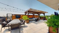 Located at 365 Terhune Rd in Princeton, NJ, this thoughtfully designed rooftop terrace offers an exceptional outdoor amenity experience across a generously proportioned open-air deck. The space is organized into several distinct functional zones, each tailored for a different style of outdoor living and entertaining.

A dedicated lounge area features teak-framed armchairs with plush cream cushions flanking built-in linear fire features, ideal for relaxed conversation beneath open skies. Overhead string lights lend a warm ambiance during evening hours. An outdoor dining zone is anchored by a large communal table surrounded by woven rope chairs, positioned adjacent to a fully equipped built-in grilling station clad in stone veneer.

A handsome wooden pergola shelters an additional seating area outfitted with sectional sofas and a wall-mounted television, creating a comfortable semi-shaded retreat. Large planter boxes filled with lush greenery and seasonal flowers are distributed throughout, adding natural warmth to the modern hardscape. A sunning area with chaise lounges provides space for relaxation, while the clean-lined white architectural façade of the rooftop service core serves as a crisp backdrop.

Perimeter railings frame sweeping panoramic views of the surrounding Princeton landscape and tree canopy, bathed in abundant natural light throughout the day.