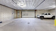 Located at 416 W Bakerview Rd in Bellingham, Washington, this single-level covered parking garage offers a well-organized and functional vehicle storage environment. The structure features a concrete construction throughout, with exposed concrete walls and support columns accented by yellow-painted protective bollards positioned at key structural points to safeguard both vehicles and columns.

The ceiling is lined with insulated paneling that spans the full extent of the space, contributing to temperature regulation within the garage. Overhead lighting is evenly distributed, ensuring clear visibility across all parking areas. The floor consists of a sealed concrete slab with clearly marked parking stalls, including designated accessible spaces identified by standard signage and pavement markings.

Vehicle access is provided via a roll-up security door, offering controlled entry and exit to the garage. A secondary personnel door is also present, providing convenient foot access. Floor drains are integrated into the slab at multiple points to manage water and debris effectively.

Along one wall, a row of individual storage units with private doors offers additional secured storage capacity for occupants. EV charging stations are also present, mounted to the walls at select stalls, reflecting modern infrastructure accommodating electric vehicle owners. Security cameras are visible overhead, providing surveillance coverage throughout the garage. The overall layout is spacious, allowing comfortable maneuverability for a variety of vehicle sizes.