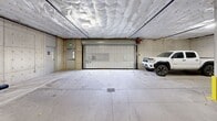 Located at 416 W Bakerview Rd in Bellingham, Washington, this single-level covered parking garage offers a well-organized and functional vehicle storage environment. The structure features a concrete construction throughout, with exposed concrete walls and support columns accented by yellow-painted protective bollards positioned at key structural points to safeguard both vehicles and columns.

The ceiling is lined with insulated paneling that spans the full extent of the space, contributing to temperature regulation within the garage. Overhead lighting is evenly distributed, ensuring clear visibility across all parking areas. The floor consists of a sealed concrete slab with clearly marked parking stalls, including designated accessible spaces identified by standard signage and pavement markings.

Vehicle access is provided via a roll-up security door, offering controlled entry and exit to the garage. A secondary personnel door is also present, providing convenient foot access. Floor drains are integrated into the slab at multiple points to manage water and debris effectively.

Along one wall, a row of individual storage units with private doors offers additional secured storage capacity for occupants. EV charging stations are also present, mounted to the walls at select stalls, reflecting modern infrastructure accommodating electric vehicle owners. Security cameras are visible overhead, providing surveillance coverage throughout the garage. The overall layout is spacious, allowing comfortable maneuverability for a variety of vehicle sizes.