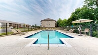 Located at 3000 Harmony Lane in Charlestown, Indiana, this outdoor recreational amenity area offers a well-appointed community pool and surrounding grounds designed for relaxation and leisure. The centerpiece is a generously sized swimming pool with striking blue tile trim, accessible via built-in entry steps and equipped with safety railings. The pool's clear, inviting water is complemented by a wide concrete sundeck that encircles the area, providing ample space for residents to move freely.

Scattered throughout the deck are multiple cushioned lounge chairs and seating arrangements, ideal for sunbathing or socializing. Market umbrellas offer shaded retreats for those seeking relief from the sun. A covered pavilion with a ceiling fan is situated adjacent to the pool, featuring brick accent details and a classic gabled roof, providing a comfortable shaded gathering space.

The entire pool area is enclosed by a sturdy wrought iron-style fence, ensuring both security and clear sightlines to the surrounding mature trees and lush greenery that frame the space. Views of the adjacent residential buildings are visible beyond the fence line, reinforcing the community-oriented setting. Safety equipment, including life rings, is stationed throughout the area. The overall atmosphere is clean, open, and welcoming, making this an attractive outdoor amenity for the community.
