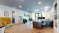 Located at 1325 14th St W in Bradenton, Florida, this thoughtfully designed single-floor space blends professional functionality with bold, contemporary style. Throughout the main areas, striking herringbone-patterned wood flooring creates a strong visual foundation, complemented by deep teal and slate-painted ceilings that add dramatic depth and character to each room.

The open office area serves as a welcoming reception and workspace, featuring a substantial dark wood desk, built-in white shelving, and an eclectic mix of vibrant artwork adorning the wainscoted walls. Colorful accent chairs in teal and yellow add personality, while recessed lighting and large glass doors flood the space with natural light and views of the lush outdoor greenery.

The living room continues the space's artistic energy with bold, large-format paintings displayed gallery-style, a comfortable sectional sofa, and elegant chandelier lighting. A set of glass doors opens to an exterior balcony, enhancing the airy, open feel.

The kitchen is clean and functional, outfitted with white shaker cabinetry, granite countertops, stainless steel appliances, a subway tile backsplash, and a peninsula with bar seating — ideal for casual gatherings. A private office nook with a built-in desk and French doors completes this well-appointed and visually dynamic space.