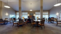 This single-floor community facility is thoughtfully designed to foster comfort and social engagement. The centerpiece is a grand, open common area distinguished by soaring vaulted ceilings and prominent structural columns that lend an architectural elegance to the space. Large windows fitted with vertical blinds and horizontal shades line the walls, allowing generous natural light to fill the room while offering views of the surrounding outdoor environment. Multiple dining tables with cushioned, striped chairs are arranged throughout, providing ample seating for group gatherings or casual meals. Pendant lighting fixtures hang from the high ceiling, complementing wall-mounted sconces that add a warm, welcoming ambiance. A dedicated lounge area features plush upholstered seating, including a bold amber sofa, a navy armchair, and a leather ottoman, creating a relaxed retreat within the larger space. A media corner includes a wall-mounted flat-screen television above a sleek dark console with decorative accents and built-in storage. The facility also includes a well-appointed kitchen equipped with dark cabinetry, an electric range, microwave, sink, and ample counter space — ideal for food preparation and catering to residents or guests. Warm-toned walls, patterned carpeting, and curated artwork throughout contribute to a cohesive, inviting atmosphere.