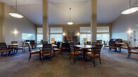 This single-floor community facility is thoughtfully designed to foster comfort and social engagement. The centerpiece is a grand, open common area distinguished by soaring vaulted ceilings and prominent structural columns that lend an architectural elegance to the space. Large windows fitted with vertical blinds and horizontal shades line the walls, allowing generous natural light to fill the room while offering views of the surrounding outdoor environment. Multiple dining tables with cushioned, striped chairs are arranged throughout, providing ample seating for group gatherings or casual meals. Pendant lighting fixtures hang from the high ceiling, complementing wall-mounted sconces that add a warm, welcoming ambiance. A dedicated lounge area features plush upholstered seating, including a bold amber sofa, a navy armchair, and a leather ottoman, creating a relaxed retreat within the larger space. A media corner includes a wall-mounted flat-screen television above a sleek dark console with decorative accents and built-in storage. The facility also includes a well-appointed kitchen equipped with dark cabinetry, an electric range, microwave, sink, and ample counter space — ideal for food preparation and catering to residents or guests. Warm-toned walls, patterned carpeting, and curated artwork throughout contribute to a cohesive, inviting atmosphere.