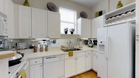 Situated in the heart of Columbus, Ohio, this single-floor residence at 250 Liberty Street offers a thoughtfully designed living space with a clean, cohesive aesthetic throughout. The kitchen serves as a standout feature, showcasing an abundance of white cabinetry that extends from floor to ceiling, providing generous storage and a bright, airy feel. White appliances, including a refrigerator with an ice and water dispenser and a dishwasher, complement the crisp, uniform palette. A double-basin sink is positioned beneath a window dressed with white blinds, allowing natural light to pour in while framing a charming windowsill with small potted plants. A mirrored backsplash runs the length of the countertops, cleverly amplifying light and adding a sense of depth to the space. Light hardwood flooring anchors the kitchen with warmth, contrasting beautifully against the white surfaces. Subtle decorative accents in yellow and olive green introduce pops of color without disrupting the clean visual flow. The overall ambiance is bright, functional, and inviting — a well-organized space that balances practicality with thoughtful design details.