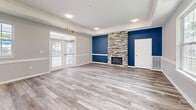 Located at 819 Forrest Dr in Newport News, Virginia, this single-floor community amenity space is thoughtfully designed to offer both comfort and functionality. The layout includes a living room, exercise room, hallway, and entrance, all finished with light-toned hardwood-style flooring that flows consistently throughout.

The living room is a standout space, anchored by a floor-to-ceiling stacked stone fireplace set against a bold navy blue accent wall. Crown molding and a tray ceiling add architectural refinement, while French doors open to an outdoor area, filling the room with natural light and providing views of the surrounding community. A wainscoting detail runs along the lower portion of the walls, contributing a clean, classic aesthetic.

Adjacent to the living room, the exercise room is well-equipped with a variety of cardio and strength-training machines, including ellipticals, a rowing machine, cable systems, and a full dumbbell rack. Large mirrors line the walls, enhancing the sense of space and supporting active use of the room. A window allows for natural light to brighten the workout environment.

The entrance and hallway are clean and functional, offering a smooth transition between areas. Overall, the space presents a bright, modern, and inviting atmosphere well-suited for community gathering and fitness activities.
