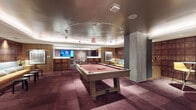 This single-floor entertainment lounge is thoughtfully designed to offer a variety of social and recreational experiences within a cohesive, upscale aesthetic. The main area is anchored by a full-size pool table set beneath a distinctive oval track lighting fixture, which draws the eye upward to the smooth, dark ceiling above. Rich mauve-toned carpet tiles cover the floor throughout, adding warmth and a sense of sophistication to the space.

A bar counter with tall barstool seating and wall-mounted flat-screen televisions serves as a central gathering point, ideal for casual viewing and socializing. Warm wood wall paneling frames multiple areas of the lounge, contributing to an inviting, club-like atmosphere. Framed artwork punctuates the walls, adding a curated touch.

A dedicated lounge alcove features a generously curved sectional sofa in amber and charcoal upholstery, paired with a round marble-top coffee table. A bold, multicolored geometric wallcovering in red, grey, and black tones energizes this nook and distinguishes it from the rest of the space. An additional seating area with a bench sofa backed by sculpted wood wall panels provides a quieter retreat.

A clean, carpeted corridor lined with recessed lighting and grey walls connects the lounge to elevators and additional amenities, completing this well-appointed entertainment destination.