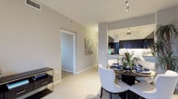This single-floor residence showcases a thoughtfully designed open-concept layout that promotes a natural flow between the dining area and kitchen. The interior is finished in a clean, neutral color palette of soft grays and warm whites, creating a bright and welcoming ambiance throughout. Light tile flooring runs through the main living areas, reflecting natural and artificial light to enhance the sense of openness.

The dining area comfortably accommodates a round dark-wood dining table surrounded by elegant upholstered white chairs, adding a refined contrast to the light surroundings. A small media console with a mirrored frame adds a stylish accent nearby. A framed piece of abstract artwork adorns the hallway wall, contributing a touch of personality to the space.

The kitchen is visible through an open archway and features dark espresso cabinetry, light countertops, and modern stainless steel appliances, including a refrigerator. Track lighting on the ceiling provides focused illumination over the kitchen workspace. A hallway leads further into the residence, with additional rooms accessible through clean, white-trimmed doorways. The overall design balances contemporary aesthetics with functional living, making it an inviting and practical space.