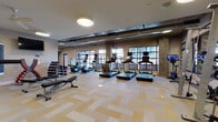 This comprehensive fitness center is thoughtfully designed across a single floor, offering a variety of dedicated workout zones to support diverse training needs. The main cardio area is bright and spacious, lined with large grid-pane windows that invite ample natural light and views of the surrounding cityscape. Multiple treadmills, stationary bikes, and elliptical machines are arranged across patterned carpet tile flooring, with wall-mounted mirrors and TVs enhancing the space. Exposed ductwork and concrete structural columns lend an industrial aesthetic throughout.

A separate strength and functional training zone features adjustable benches, a heavy bag, TRX-style suspension trainers on an orange rig, and a full Life Fitness cable machine — all complemented by a ceiling fan, mounted flatscreen TV, and cubbied storage wall. An additional dedicated cycling studio offers wood-look flooring, multiple spin bikes, large framed mirrors, and pendant lighting for an immersive class environment.

Two distinct studio rooms with warm hardwood-style floors serve yoga, stretching, and group fitness classes. One includes a large display screen, exercise balls, and yoga blocks, while glass partitions allow visual connectivity to the main floor. The overall ambiance is modern, clean, and energizing — built to accommodate a full spectrum of fitness activities in one cohesive facility.