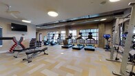This comprehensive fitness center is thoughtfully designed across a single floor, offering a variety of dedicated workout zones to support diverse training needs. The main cardio area is bright and spacious, lined with large grid-pane windows that invite ample natural light and views of the surrounding cityscape. Multiple treadmills, stationary bikes, and elliptical machines are arranged across patterned carpet tile flooring, with wall-mounted mirrors and TVs enhancing the space. Exposed ductwork and concrete structural columns lend an industrial aesthetic throughout.

A separate strength and functional training zone features adjustable benches, a heavy bag, TRX-style suspension trainers on an orange rig, and a full Life Fitness cable machine — all complemented by a ceiling fan, mounted flatscreen TV, and cubbied storage wall. An additional dedicated cycling studio offers wood-look flooring, multiple spin bikes, large framed mirrors, and pendant lighting for an immersive class environment.

Two distinct studio rooms with warm hardwood-style floors serve yoga, stretching, and group fitness classes. One includes a large display screen, exercise balls, and yoga blocks, while glass partitions allow visual connectivity to the main floor. The overall ambiance is modern, clean, and energizing — built to accommodate a full spectrum of fitness activities in one cohesive facility.