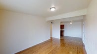 This single-floor space offers a clean, functional layout with a cohesive design throughout. The living area is finished with warm, honey-toned hardwood floors that extend seamlessly into the adjoining spaces, adding character and a sense of continuity. Crisp white walls reflect light throughout the interior, contributing to an open and airy atmosphere.

A structural beam detail defines the transition between the main living area and the kitchen zone, providing subtle architectural interest without interrupting the open flow of the space. The kitchen features rich, dark wood cabinetry that provides a warm contrast to the lighter tones of the surrounding walls and flooring. A ceiling fan in the kitchen area ensures comfortable airflow and practical ventilation.

Flush-mount ceiling lighting is present in the living area, providing straightforward, reliable illumination. The layout is efficiently designed, with a doorway connecting the main living space to additional rooms, allowing for easy movement throughout the unit. Overall, the space combines practical finishes with a warm, welcoming ambiance, making it well-suited for everyday living.