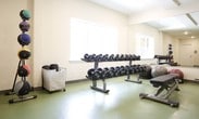 This fitness center is a comprehensively outfitted single-floor facility designed to support a full range of workout routines. The space is organized into distinct workout zones, each tailored to a specific type of exercise. The cardio area houses multiple treadmills, each paired with its own wall-mounted flat-screen television, allowing users to stay entertained during longer sessions. Alongside the treadmills, a selection of elliptical trainers and stationary bikes provides varied low-impact cardiovascular options. A large wall mirror spans one side of the room, visually expanding the space and allowing users to monitor their form. The free weights section features a well-stocked dumbbell rack with a broad range of weights, accompanied by stability and exercise balls for functional training. Natural light filters in through multiple windows that overlook a green outdoor area, creating a bright and inviting atmosphere. The flooring throughout is a resilient, easy-to-clean green material well-suited to high-traffic fitness use. Recessed lighting supplements the natural light, ensuring the space remains well-lit at all hours. A water dispenser and waste receptacle are conveniently positioned within the space, and glass-panel entry doors contribute to the open, accessible feel of the facility.