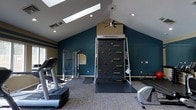 This single-floor fitness center offers a thoughtfully designed workout space that combines functionality with an inviting atmosphere. The room's most striking architectural feature is its vaulted ceiling, which rises to a peak and is fitted with skylights that draw in natural light, enhancing the sense of openness. A ceiling fan ensures comfortable air circulation during workouts.

The space is anchored by a standout Treadwall — a motorized vertical climbing wall equipped with colorful handholds — serving as both a functional and visually dynamic centerpiece. Complementing this unique piece of equipment are a treadmill, an elliptical machine, a cable resistance training station, and additional strength and flexibility training equipment, providing a comprehensive full-body workout experience.

The interior is finished with deep teal accent walls offset by neutral upper walls and trim, giving the space a modern and motivating aesthetic. Recessed lighting supplements the natural light from the skylights, ensuring the room is well-lit at all times. The floor is covered with a durable, patterned carpet with protective rubber mats placed beneath the equipment. A window on one wall adds further natural light and ventilation, rounding out this versatile and well-appointed fitness facility.