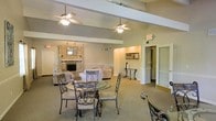 This single-floor space offers an open, welcoming layout that seamlessly combines a dining area and a lounge. The room is anchored by a striking stone fireplace, which serves as a focal point and adds a warm, classic character to the overall design. Vaulted ceilings with exposed beams lend a sense of spaciousness and architectural interest, while two ceiling fans ensure comfortable airflow throughout the area. The dining area features an elegant glass-top table with ornate wrought-iron chairs, providing a refined setting for gatherings. Adjacent to it, a plush seating arrangement with neutral-toned sofas and chairs creates a relaxed lounge environment ideal for conversation or quiet relaxation. The neutral color palette of beige and cream tones throughout the walls, carpet, and furnishings gives the space a cohesive, understated elegance. A hallway visible at the rear leads to additional rooms, suggesting further utility beyond the main gathering area. Framed artwork adorns the walls, adding subtle decorative touches. The combination of architectural detailing, cozy fireplace, and thoughtfully arranged furnishings makes this a functional and inviting communal environment.