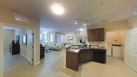 This single-story residence is designed around an open-concept floor plan that seamlessly connects the kitchen, dining, and living areas into one cohesive, flowing space. The kitchen serves as a central hub, featuring dark espresso cabinetry, granite countertops with a deep, richly veined finish, and a wraparound peninsula that doubles as a casual gathering point. Black appliances complement the darker cabinetry, while track lighting overhead provides focused illumination across the workspace.

The living and dining areas extend naturally from the kitchen, unified by light-toned tile flooring that runs consistently throughout the space, contributing to a bright and airy atmosphere. Large windows dressed with vertical blinds invite abundant natural light, enhancing the warm beige tones of the walls. The dining area is anchored by a glass-top table surrounded by upholstered chairs, with a decorative framed mirror mounted on the wall adding visual depth. The living area offers a comfortable, neutral-toned seating arrangement beneath a ceiling fan, creating a relaxed and welcoming environment. A hallway visible from the main living space suggests additional private rooms beyond, completing this well-appointed, functionally designed residence.