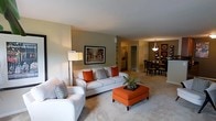 This single-floor space showcases an open-concept layout that seamlessly connects the living, dining, and kitchen areas, creating a cohesive and functional environment. The living area is anchored by a plush white sofa and accent chair, both complemented by bold orange and patterned throw pillows that add a vibrant pop of color against the neutral, warm-toned walls. An orange upholstered ottoman serves as a central coffee table, reinforcing the room's color palette. A floor lamp provides warm ambient lighting, while a framed piece of colorful artwork adorns the feature wall, adding visual interest. The space is finished with soft, light-toned carpeting throughout, contributing to a comfortable and inviting atmosphere. Beyond the living area, the dining space is furnished with dark wood chairs surrounding a dining table, lit by an elegant pendant light fixture overhead. Wall décor accents the dining space, adding to its polished look. The kitchen is partially visible in the background, featuring light cabinetry and a hallway leading further into the home. Overall, the space presents a clean, well-coordinated aesthetic with a warm, welcoming ambiance.