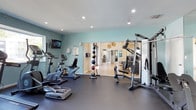 This fitness center offers a thoughtfully designed workout space finished with soft teal walls and a clean white ceiling outfitted with recessed lighting. The dark rubberized flooring provides a durable, slip-resistant surface suited for a variety of exercise activities. Cardio equipment, including stationary bikes and a Nautilus machine, is arranged along one side of the room, complemented by a wall-mounted flat-screen television for entertainment during workouts. A vertical medicine ball rack provides convenient access to functional training tools. On the opposite side, a multi-station cable strength training machine offers versatile resistance options. Large glass sliding doors open directly into an adjacent room, filling the fitness area with natural light and creating a seamless indoor connection. The neighboring space, visible through the doors, features tile flooring, artwork, and additional seating, suggesting a lounge or common area. The overall layout is open and uncluttered, allowing for comfortable movement throughout the space. The combination of natural light, modern equipment, and a clean color palette contributes to an inviting and functional environment well-suited for daily exercise routines.