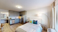 Located at 2070 McMillan Ave in North Charleston, SC, this thoughtfully designed unit maximizes every inch of its compact footprint. The open-concept layout seamlessly integrates the sleeping area with a fully equipped kitchen, creating a functional and cohesive living space. The kitchen features stainless steel appliances — including a refrigerator, microwave, and dishwasher — complemented by gray flat-front cabinetry that extends to the ceiling for ample storage and a white subway tile backsplash with an undermount sink.

The sleeping area is bright and airy, with light wood-look flooring that flows throughout the entire unit, white walls, and a large window that allows natural light to fill the room. A built-in wall shelving system with a dedicated desk area provides a practical workspace, making the unit well-suited for remote work or study.

A vestibule area near the entry serves as a transitional space, offering a closet with hanging rod and overhead shelving alongside a granite-topped vanity counter. The full bathroom is cleanly finished with a granite vanity, framed mirror, and a tub/shower combination with a curtain enclosure, all set against light wood flooring and crisp white walls. The unit's efficient design, modern finishes, and practical storage solutions make it a well-appointed and functional living space.