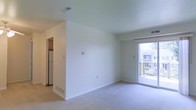 This single-floor residence features a clean, minimalist layout designed to maximize usability and comfort. The main living area is finished with neutral wall-to-wall carpeting and crisp white walls, creating a bright and airy atmosphere. A sliding glass door dominates one wall, offering direct access to a private outdoor balcony and allowing generous natural light to fill the space. The view through the glass door reveals a wood-decked balcony with neighboring building views beyond.

A partial wall divides the living area from the kitchen, which is visible in the background and features a stainless steel refrigerator and what appears to be a window treatment above. The kitchen connection provides an open yet defined separation between the living and cooking spaces.

The unit includes flush-mounted ceiling vents for climate control, and a wall-mounted thermostat is visible in the living area. Baseboards throughout are white and well-maintained, consistent with the overall clean aesthetic. The space benefits from a simple, functional design that offers a comfortable canvas ready for personalization.