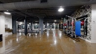Located at 300 N Sixth Ave in Minneapolis, this comprehensive fitness facility occupies a generously sized single floor with a distinctly industrial aesthetic. Exposed concrete ceilings with visible ductwork, large structural columns, and polished concrete floors define the main training areas, lending the space a raw, urban character. The primary workout floor houses a full array of cardio machines — including treadmills, ellipticals, and stationary bikes — alongside strength training equipment, all arranged beneath pendant and recessed lighting reflected in the gleaming floors. A dedicated boxing area features a full-size boxing ring, heavy punching bags suspended from overhead racks, and open floor space for combat fitness training. A separate strength and functional training zone provides rubber-floored space lined with wall-to-wall mirrors and well-stocked racks of kettlebells and dumbbells in a range of sizes. The facility also includes a bright group fitness studio with large industrial-style windows that invite natural light, rubber flooring, and floor-to-ceiling mirrors. An additional studio with warm hardwood flooring, a ballet barre, and generous window views offers a refined space for barre or dance-based classes. Black-framed glass partitions provide visual separation between zones while maintaining an open, connected feel throughout.