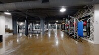 Located at 300 N Sixth Ave in Minneapolis, this comprehensive fitness facility occupies a generously sized single floor with a distinctly industrial aesthetic. Exposed concrete ceilings with visible ductwork, large structural columns, and polished concrete floors define the main training areas, lending the space a raw, urban character. The primary workout floor houses a full array of cardio machines — including treadmills, ellipticals, and stationary bikes — alongside strength training equipment, all arranged beneath pendant and recessed lighting reflected in the gleaming floors. A dedicated boxing area features a full-size boxing ring, heavy punching bags suspended from overhead racks, and open floor space for combat fitness training. A separate strength and functional training zone provides rubber-floored space lined with wall-to-wall mirrors and well-stocked racks of kettlebells and dumbbells in a range of sizes. The facility also includes a bright group fitness studio with large industrial-style windows that invite natural light, rubber flooring, and floor-to-ceiling mirrors. An additional studio with warm hardwood flooring, a ballet barre, and generous window views offers a refined space for barre or dance-based classes. Black-framed glass partitions provide visual separation between zones while maintaining an open, connected feel throughout.