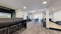 Located at 425 Ballantrae Drive in Winnipeg, this single-floor amenity lounge offers a well-rounded entertainment and social space designed with a contemporary aesthetic. The interior features light wood-effect flooring, clean white walls, and recessed pot lighting complemented by a striking exposed-bulb chandelier that serves as a central design focal point in the main gathering area.

The space is thoughtfully divided into several activity zones. A dedicated golf simulator room provides an immersive experience complete with a large projection screen, a service counter with bar stools, and lounge seating. Adjacent to this, a row of classic arcade cabinets lines one wall, adding a retro gaming element to the modern setting.

The central lounge area is furnished with multiple bar-height tables and stools, creating a casual and social atmosphere ideal for gathering. Large windows along one wall allow natural light to fill the space and provide views to the exterior. A shuffleboard table and a billiards table are also present, rounding out the recreational offerings.

A built-in counter area with bench seating provides additional casual seating, and wall-mounted displays are positioned throughout. The overall design balances functionality with a sleek, urban sensibility, making this a versatile amenity space suited for both relaxed socializing and active recreation.