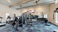 This fitness center in Fort Worth, Texas offers a thoughtfully designed, single-floor exercise space accommodating a comprehensive range of workout options. The room features soaring ceilings with exposed decorative beams and dual ceiling fans that promote airflow and comfort during workouts. Large windows along the walls flood the space with natural light, providing views of the surrounding outdoor area and creating an open, energizing atmosphere.

The exercise room is outfitted with a robust selection of equipment, including multiple treadmills, elliptical cross-trainers, and strength machines from brands such as TRUE Fitness and Vesta. A multi-functional cable machine anchors the strength training zone, while a dedicated TRX suspension training station and a well-stocked rack of medicine balls, kettlebells, and resistance accessories support functional fitness routines. A full-length wall mirror expands the sense of space and allows for form-checking during exercises. A wall-mounted flat-screen television adds an entertainment element to the environment.

The flooring consists of stylish modular carpet tiles in a blue and teal patterned design, adding visual interest while providing cushioning underfoot. Recessed lighting complements the natural light, ensuring the space remains bright throughout the day. The overall ambiance is modern, clean, and motivating — well-suited for a wide range of fitness levels and training styles.