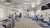This fitness center offers a spacious, single-floor layout thoughtfully designed to accommodate a comprehensive range of workout activities. The interior features elevated tray ceilings with crown molding, recessed panel lighting, and ceiling fans, all contributing to a bright and comfortable exercise environment. Large arched windows along the walls invite abundant natural light, creating an open and energizing atmosphere throughout the space.

The main workout area is carpeted and divided into distinct functional zones. The cardio section is lined with multiple treadmills and elliptical machines, positioned to take advantage of the natural light from the arched windows. Adjacent to this, a strength-training zone houses a variety of cable machines and resistance equipment, along with a dedicated free-weight area featuring a full dumbbell rack and wall-mounted mirrors for form checks. Flat and adjustable benches, a pull-up and dip station, and rubber floor mats round out the strength section.

A separate functional training area is outfitted with medicine balls, stability balls, and wall-mounted accessories, catering to flexibility and balance-focused workouts. Framed instructional charts on the walls provide exercise guidance. French doors and a glass-panel entry door connect the space, while a mounted television keeps members informed or entertained during their sessions. The overall design is clean, neutral, and well-maintained, offering a professional and welcoming fitness environment.