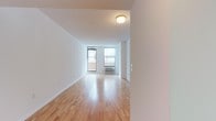This single-floor space offers a clean, minimalist aesthetic defined by crisp white walls and warm, light-toned hardwood flooring that reflects natural light throughout. The open layout creates a welcoming sense of spaciousness, making the area feel airy and versatile. Large windows along the far wall frame views of the surrounding exterior, flooding the interior with abundant daylight and contributing to the bright, open atmosphere. A wall-mounted HVAC unit positioned beneath the windows ensures year-round climate control. The smooth, neutral surfaces throughout provide a blank canvas adaptable to a variety of uses and furnishing styles. Ceiling-mounted flush lighting complements the natural illumination, ensuring the space remains well-lit at all hours. The textured ceiling adds a subtle visual detail that contrasts gently with the otherwise streamlined interior. The combination of natural light, hardwood floors, and clean architectural lines gives this space a polished yet approachable character, ideal for those seeking a functional and well-maintained environment with a timeless, understated design sensibility.