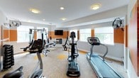 This fitness center offers a comprehensive workout environment designed to accommodate a wide range of exercise needs. The space is defined by a bold, energizing aesthetic — warm orange accent bands run along the walls at mid-height, contrasting with neutral cream tones above and below, and complemented by vibrant abstract artwork throughout.

The main workout area is generously sized and well-lit, benefiting from multiple large windows that invite natural light and views of surrounding greenery. Flush-mount and recessed ceiling fixtures provide consistent overhead lighting throughout. The flooring features a combination of neutral carpet tiles with orange accent squares, reinforcing the facility's signature color palette.

Cardio equipment includes treadmills, elliptical trainers, and a stationary cycle, positioned to allow comfortable spacing between users. A multi-station cable and weight machine serves as the centerpiece for strength training, accompanied by a rack of dumbbells for free weight exercises. A wall-mounted flat-screen television adds an entertainment element to the workout experience.

A dedicated "Stretch" area is accessible through glass-paned doors, offering a quieter zone for flexibility work and cool-downs. The facility also features a wall-mounted fan for additional ventilation and hand sanitizer stations. The overall design is clean, functional, and motivating — built to support an active lifestyle.