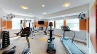 This fitness center offers a comprehensive workout environment designed to accommodate a wide range of exercise needs. The space is defined by a bold, energizing aesthetic — warm orange accent bands run along the walls at mid-height, contrasting with neutral cream tones above and below, and complemented by vibrant abstract artwork throughout.

The main workout area is generously sized and well-lit, benefiting from multiple large windows that invite natural light and views of surrounding greenery. Flush-mount and recessed ceiling fixtures provide consistent overhead lighting throughout. The flooring features a combination of neutral carpet tiles with orange accent squares, reinforcing the facility's signature color palette.

Cardio equipment includes treadmills, elliptical trainers, and a stationary cycle, positioned to allow comfortable spacing between users. A multi-station cable and weight machine serves as the centerpiece for strength training, accompanied by a rack of dumbbells for free weight exercises. A wall-mounted flat-screen television adds an entertainment element to the workout experience.

A dedicated "Stretch" area is accessible through glass-paned doors, offering a quieter zone for flexibility work and cool-downs. The facility also features a wall-mounted fan for additional ventilation and hand sanitizer stations. The overall design is clean, functional, and motivating — built to support an active lifestyle.