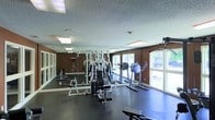This single-floor fitness center is designed to support a comprehensive workout experience. The space is outfitted with a multi-station strength training machine, free-weight benches, and a selection of cardio equipment, including an elliptical and a stationary bike, providing users with a variety of exercise options. Dark rubber flooring covers the workout area, offering durability and impact absorption underfoot. A full wall of mirrors along one side visually expands the room while allowing exercisers to monitor their form during workouts. The textured ceiling features recessed fluorescent lighting, ensuring the space is well-lit throughout. Large windows along the exterior wall allow natural light to fill the room and offer views of the greenery outside, contributing to an open, motivating atmosphere. The warm brown wall tones complement the dark flooring, creating a focused and energetic environment. Overall, the fitness center is thoughtfully arranged to maximize usable floor space while accommodating both strength and cardio training needs.