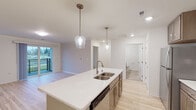 This single-floor residence offers a thoughtfully designed open-concept layout with modern finishes throughout. The kitchen serves as a central hub, anchored by a large island with a white quartz countertop, a double stainless steel sink, and built-in dishwasher. Taupe-toned cabinetry lines the walls alongside stainless steel appliances, including a range, microwave, and refrigerator, all illuminated by elegant glass pendant lights. The kitchen flows seamlessly into a bright living area, which features light hardwood flooring and a sliding glass door opening to an outdoor balcony with views of a green landscape.

The bedrooms are well-proportioned and filled with natural light from large windows. One notable bedroom features vaulted ceilings accented by a ceiling fan, creating an airy, open atmosphere with outdoor views. Additional bedrooms are finished with the same light hardwood flooring and neutral walls.

The bathroom is clean and functional, featuring warm-toned tile flooring, a single vanity with light wood cabinetry, a white countertop, and a large mirror. Recessed lighting and ample ventilation are present throughout. The consistent use of light tones, hardwood floors, and modern fixtures gives the entire space a cohesive, contemporary aesthetic.