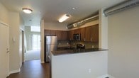 This two-floor space showcases a modern, industrial-influenced design that blends functionality with contemporary style. The kitchen serves as a central feature, outfitted with rich, warm-toned wood cabinetry that extends to the upper walls, providing generous storage throughout. Dark granite countertops run along the workspace and continue onto a peninsula that opens toward the living area, creating a natural flow between cooking and entertaining spaces. Stainless steel appliances, including a full-size refrigerator, built-in microwave, and range, complement the kitchen's polished aesthetic. An exposed metallic ventilation duct runs prominently overhead, lending an urban, loft-like character to the space. Dark hardwood flooring grounds the interior and adds warmth, contrasting effectively with the light-toned walls. Natural light filters in through a large window visible from the kitchen, brightening the adjacent living area. The open-concept layout between the kitchen and living space enhances the overall sense of roominess. The two-floor configuration offers additional living or sleeping space, making this a versatile and well-appointed residence with a distinctly modern edge.