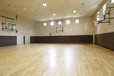 Located in Woodstock, Georgia, this single-floor indoor basketball court offers a well-appointed athletic space designed for active recreation. The court's most prominent feature is its expansive hardwood floor, which provides a smooth, durable surface suited for basketball and a variety of other athletic activities.

The soaring ceiling height creates an open, airy atmosphere essential for unobstructed play. A wall-mounted basketball system with a glass backboard and standard hoop is positioned at one end of the court, while wall-mounted hardware on the opposite end suggests the option for a second hoop. Dark padded wall coverings line the lower portion of all walls, providing important safety protection for players during active use.

Natural light enters the space through rows of clerestory windows positioned high along the walls, supplemented by overhead fluorescent lighting to ensure consistent illumination throughout. On one side of the court, large interior windows offer a view into an adjacent fitness or weight room area, and two standard doors provide access to additional rooms or storage areas. The neutral tan upper walls and dark lower padding create a clean, functional aesthetic throughout the space.