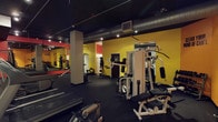 This single-floor fitness facility offers a comprehensive workout environment designed to accommodate a variety of training needs. The space is immediately defined by its striking interior palette — vivid yellow walls contrast sharply against a dark, exposed black ceiling, creating an energetic and motivating atmosphere. Industrial-style ductwork and track lighting run overhead, providing focused illumination throughout the workout floor.

The space is equipped with a diverse selection of strength training machines, including a multi-function cable and weight stack system, an adjustable bench press station, and various resistance machines suited for both upper and lower body workouts. A dedicated free weights area lines one wall, with a full-length mirror allowing for form checks during exercises. Dumbbells and weight racks are neatly arranged alongside cardio equipment, including treadmills visible toward the front of the space.

Rubber flooring covers the entire workout area, providing durability and safety underfoot. Structural columns are integrated into the layout, subtly dividing the floor into functional training zones without disrupting the open feel. The overall design is utilitarian yet purposeful, offering a no-frills, high-function environment built entirely around athletic performance and training efficiency.