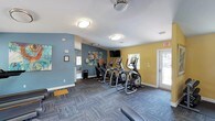 Located at 711 Underwood Ave. in Pensacola, Florida, this fitness center offers a well-equipped and inviting space designed to support an active lifestyle. The single-floor facility is finished with a bold two-tone color scheme, pairing cool slate-blue accent walls with warm yellow surfaces to create an energizing atmosphere throughout.

The main exercise area is generously sized and features a row of modern elliptical machines and cardio equipment, providing ample room for comfortable workouts. Underfoot, durable patterned carpet tiles cover the floor, offering both cushioning and easy maintenance. Flush-mounted ceiling lights ensure consistent, even illumination across the space.

A wall-mounted flat-screen television is positioned for easy viewing during workouts, adding a convenient entertainment option. Natural light filters in through multiple windows and a glass-panel exterior door, contributing to an open and airy feel. The exit door provides direct outdoor access, enhancing ventilation and convenience.

Decorative artwork adorns the walls, lending a welcoming, community-oriented character to the space. Additional amenities such as a wall clock and safety signage reflect thoughtful attention to functionality. Overall, this fitness center combines practical equipment, vibrant design, and natural light to deliver a motivating environment for residents and users alike.