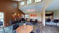 This single-floor gathering space combines a café-style atmosphere with thoughtful architectural design, making it an inviting destination for socializing or casual meetings. The room is anchored by dramatically high, vaulted ceilings that amplify the sense of openness, while a row of clerestory windows near the roofline floods the upper walls with natural light and treetop views, enhancing the airy feel throughout.

The interior features a rich contrast of warm earth tones — deep chocolate-brown accent walls complement the cream-colored upper walls — creating a cozy yet dynamic ambiance. Colorful abstract artwork in red, orange, and gold tones adorns the walls, adding a vibrant artistic character to the space. Patterned, multi-tone carpeting in neutral earth tones grounds the room while adding visual texture underfoot.

Furnishings consist of several round wooden bistro-style tables paired with sleek, dark side chairs, thoughtfully arranged to encourage conversation. A built-in counter along one wall provides additional seating or workspace. Track lighting and pendant fixtures supplement the natural light, ensuring the space remains well-lit at any time of day. A connected area, visible through an opening, suggests additional amenities nearby, rounding out this well-appointed communal space.
