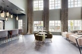 Located at 3110 Doe River Way in Matthews, NC, this elegantly designed living space delivers a striking first impression through its dramatic double-height ceilings and expansive floor-to-ceiling windows framed by floor-length velvet drapes. The open-concept layout is anchored by large-format herringbone tile flooring that flows seamlessly throughout, lending a refined sense of continuity. An eye-catching abstract mural serves as a bold focal point on one wall, complementing the curated art pieces displayed throughout the space. A sleek industrial-style staircase with metal railings and warm wood treads leads to an upper mezzanine level, adding vertical dimension and architectural interest. The lounge area features plush velvet seating in earthy olive tones arranged around gold-accented tables, while a sectional sofa anchors a relaxed seating nook beneath large-scale artwork. A bar counter with upholstered stools, illuminated by sculptural pendant lighting, creates an inviting social hub. Natural light pours in from multiple oversized windows, offering views of the surrounding exterior. The overall ambiance blends contemporary sophistication with warm, layered textures, resulting in a space that is both visually dynamic and functionally inviting.