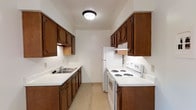 This single-floor space offers a practical and well-organized layout designed for comfortable everyday living. The galley-style kitchen is a standout feature, thoughtfully arranged to maximize efficiency and storage. Warm honey-toned wood cabinetry lines both sides of the kitchen from floor to ceiling, providing generous storage space for cookware, pantry items, and more. The countertops offer ample workspace and are complemented by a double-basin sink positioned conveniently along one wall. A white electric coil range and a full-size refrigerator round out the kitchen's appliance suite, delivering all the essentials for home cooking. Overhead, a flush-mount ceiling light provides steady, even illumination throughout the kitchen corridor. The rich, dark hardwood-style flooring adds warmth and contrast to the lighter cabinetry and white appliances, lending the space a cozy and grounded feel. At the far end of the kitchen, a wooden door leads to an adjacent area, hinting at a connected and functional floor plan. Overall, the space presents a clean, no-frills aesthetic that prioritizes utility and warmth in equal measure.
