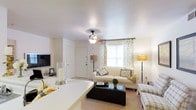 This single-floor residence is designed with an open-concept layout that seamlessly connects the living, dining, and kitchen areas into one cohesive and functional space. The living room is comfortably proportioned, featuring plush wall-to-wall carpeting, white walls, and a ceiling fan with a light fixture that provides both comfort and illumination. A large window dressed with patterned curtains allows natural light to fill the space, enhancing the bright and airy atmosphere. The room accommodates a sofa, accent chairs, and a dark coffee table, creating a well-balanced seating arrangement.

Adjacent to the living area, the dining space features a compact round table with chairs, accented by wall sconces and framed artwork that add warmth to the neutral palette. A half-wall partition separates the kitchen, which includes overhead cabinetry, a countertop pass-through, and a sink, maintaining visual connectivity while defining the cooking area. Throughout the space, the consistent use of light-colored walls, soft carpeting, and understated décor contributes to a clean, cohesive, and inviting ambiance suited for everyday living.