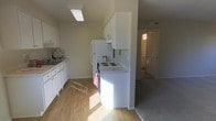 This single-floor space includes a well-organized kitchen outfitted with white cabinetry, a white refrigerator, and an electric coil stove. The kitchen features a clean, compact layout with ample upper and lower cabinet storage, a sink along one wall, and light wood-style flooring that adds warmth to the space. A ceiling-mounted light fixture provides overhead illumination, while natural light filters in from an adjacent area, brightening the kitchen. The countertops are white, maintaining a cohesive, neutral color palette throughout. A partial wall separates the kitchen from the adjoining carpeted area, which transitions into what appears to be a hallway or additional living space beyond. A doorway visible in the background hints at further rooms, with natural light suggesting a window nearby. The neutral wall tones and light flooring contribute to a bright, airy atmosphere throughout the space. Overall, the layout is practical and straightforward, with a clear flow between the kitchen and the adjoining areas.