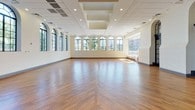 The Jefferson Ballroom is a distinguished event venue that seamlessly blends classic architectural elegance with modern functionality. The expansive main hall is defined by its warm, richly toned hardwood floors laid in a chevron pattern, creating a visually dynamic foundation that anchors the entire space.

Lining the walls are a series of tall, arched windows framed in bold dark steel, allowing abundant natural light to flood the interior while offering views of lush surrounding greenery. These architectural details lend the ballroom a timeless, sophisticated character. Along one wall, a row of arched doorways with matching dark-framed glass doors provides access to adjacent areas, maintaining the cohesive design language throughout.

The ceiling is a notable feature, incorporating a stepped coffered detail at the center with recessed lighting, adding depth and architectural interest above. Supplementary recessed lighting is distributed evenly across the ceiling, ensuring the space is well-lit for any occasion.

The open floor plan offers exceptional flexibility, making the Jefferson Ballroom well-suited for receptions, galas, corporate events, and more. The neutral white walls serve as a versatile backdrop, adaptable to a wide range of décor and event styles. Overall, the space exudes an atmosphere of refined grandeur, combining classic design elements with a clean, contemporary finish.