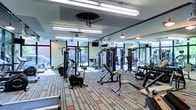 This fitness center offers a thoughtfully designed workout environment built to accommodate a variety of exercise needs. The space is outfitted with an extensive selection of strength-training machines, cable systems, and a dedicated free weights section complete with a dumbbell rack, providing users with comprehensive options for resistance training. Cardio equipment, including a recumbent bike, is also available for low-impact exercise. The interior is bright and welcoming, with large floor-to-ceiling windows along multiple walls that flood the space with abundant natural light and offer views of the surrounding outdoor environment. Full-length mirrors line one wall, visually expanding the room while allowing users to monitor their form during workouts. The ceiling features a combination of industrial-style pendant lights and fluorescent strip lighting, ensuring consistent illumination throughout. A ceiling-mounted television provides entertainment during workouts. The floor is covered in a distinctive geometric-patterned carpet in neutral tones accented with red, adding a stylish and functional touch to the overall aesthetic. The layout is open and organized, maximizing usable floor space and ensuring comfortable movement between equipment stations.